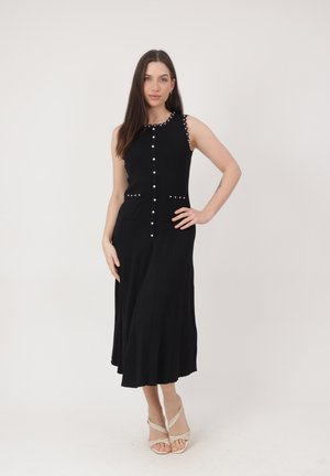 Donna con un vestito midi nero senza maniche con bottoni di perla e tacchi bianchi con cinturini, in piedi con una mano sui fianchi davanti a uno sfondo semplice.