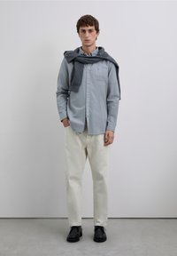 Lyseblå skjorte med knapper parret med beige bukser og sorte sko. En mørkegrå sweater er lagt over skuldrene. Enkel design, rene linjer.