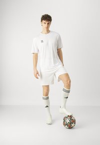 Weißes Fußballtrikot bestehend aus einem Kurzarmshirt und Shorts. Strukturiertes Material, kombiniert mit Fußballsocken mit drei schwarzen Streifen und Stollenschuhen. Ball zu Füßen.
