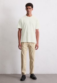 Hellgelbes T-Shirt mit kurzen Ärmeln, kombiniert mit beigen Hosen und schwarzen Slippern, vor einem einfarbigen Hintergrund stehend.