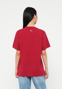 Persoană cu păr lung și negru, îmbrăcată cu un tricou roșu larg pe care este inscripționat textul "Hugo" pe spate, purtând blugi de culoare albastru deschis, întoarsă cu fața. 