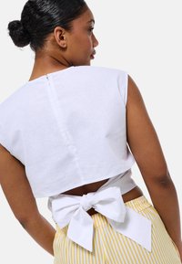 Haut court blanc avec un détail de nœud au dos, des manches courtes et une fermeture éclair discrète. Assorti avec un pantalon rayé jaune et blanc.