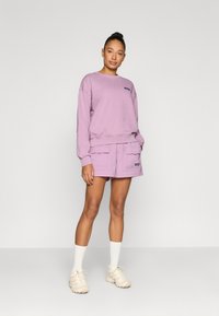 Lys lilla genser med ribbestrikkede mansjetter og kant, kombinert med matchende shorts med sidelommer og merkevare. Brukt med hvite sokker og joggesko.