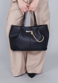 Person i beiga byxor som håller en stor svart texturerad handväska med guldkedja och låsdetalj, iklädd svarta spetsiga skor.