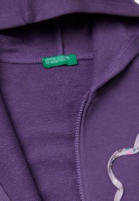 Felpa zip con cappuccio di colore viola, realizzata in tessuto texture, fodera del cappuccio con un motivo floreale e etichetta verde "UNITED COLORS OF BENETTON."