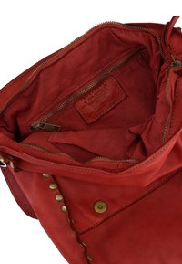 Czerwona skórzana torebka z mosiężnymi ćwiekami, otwarta u góry, ukazująca wewnętrzną kieszeń na zamek i skórzaną metkę z logo wewnątrz.