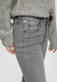 Jean gris en denim, avec une coupe slim, un design à cinq poches et des boutons foncés en accent, associé à un pull en tricot gris clair.