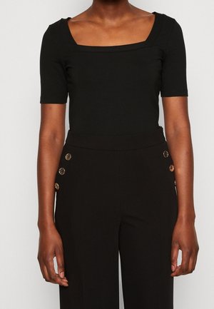 Vrouw draagt een zwart topje met een vierkante hals en korte mouwen, en een zwarte broek met hoge taille met aan elke kant drie gouden knopen.