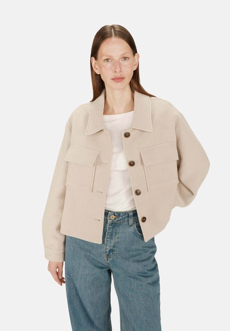 Kvinde med glat brunt hår iført beige knapjakke med to store forlommer, hvid top og blå jeans foran en ensfarvet baggrund.