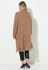 Cardigan en tricot beige avec des touches multicolores, manches longues, ouverture sur le devant et détails en franges à l'ourlet ; associé à un pantalon noir ajusté.
