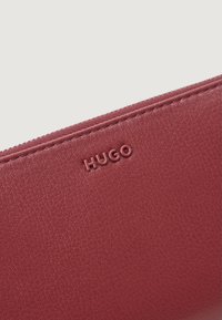 Cartera roja de cuero con el logo 'HUGO' en relieve. Presenta una superficie texturizada y una costura a lo largo del cierre de cremallera.