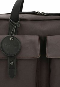 Sac en tissu marron foncé avec deux poches avant à rabat et une poignée en cuir noir, doté d'une étiquette ronde en cuir noir avec le nom de la marque embossé.