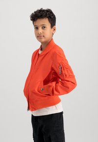 Orange bomber jacket with ribbed collar and cuffs, front zipper, side pockets, and a zippered sleeve pocket. Styled over a white shirt.
