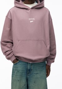 Pullover-Hoodie in Lavendel mit einer Fronttasche, versehen mit weißem Logo-Text. Hergestellt aus weichem Material mit lockerer Passform und gerippten Bündchen.