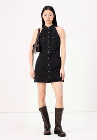 Zwart denim mouwloos jurkje met knoopsluiting aan de voorkant, kraag, twee borstzakken en een elastische taille. G gecombineerd met bruine kniehoge laarzen.