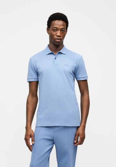 Homme portant un polo à manches courtes bleu clair et un pantalon assorti, debout devant un fond blanc uni.