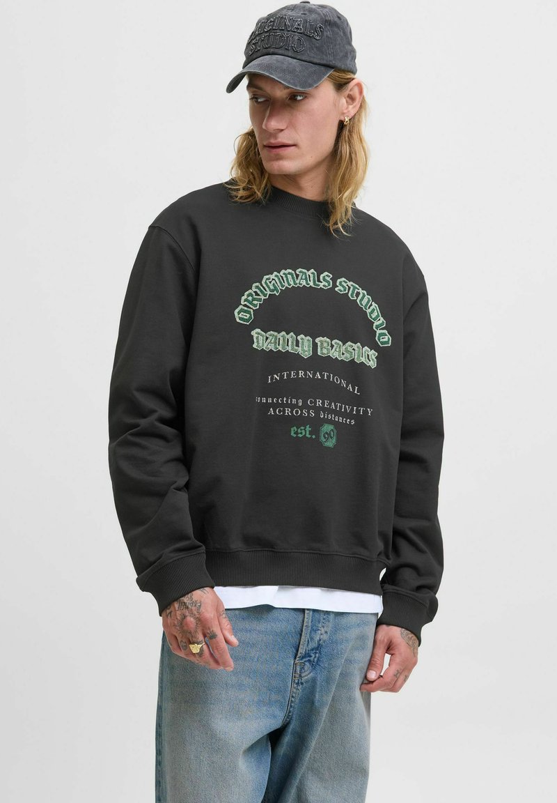 Ung mand med langt hår iført en sort sweatshirt med grøn tekst, blå jeans, en mørk kasket og synlige håndtatoveringer.