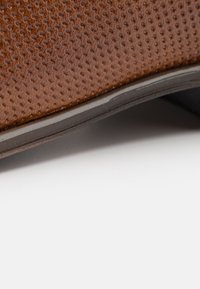 Bruna läderskor med en strukturerad yta som har små perforeringar. Sulan är lätt svängd. Inga ytterligare dekorationer synliga.