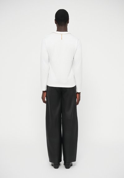 Biely dlhý rukáv s detailom reťaze na výstrihu, v kombinácii so širokými čiernymi nohavicami, z pohľadu zozadu. Hladká textúra látky.