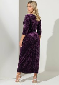 Mujer con cabello rubio, vestida con un largo vestido de terciopelo morado y sandalias de tacón plateadas, de pie y mirando por encima del hombro.