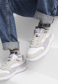 Zapatillas blancas y gris claro con texturas de ante y malla, con acentos morados y un logotipo visible. Combínalas con unos jeans oscuros y calcetines estampados.