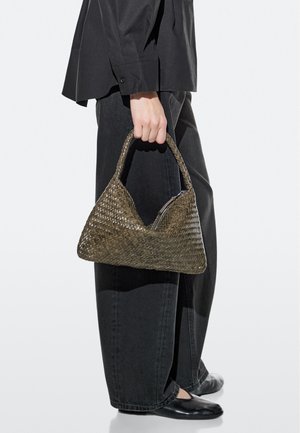 Mano che tiene una borsa intrecciata verde oliva con cerniera, indossando pantaloni larghi neri, camicia nera e scarpe piatte nere.