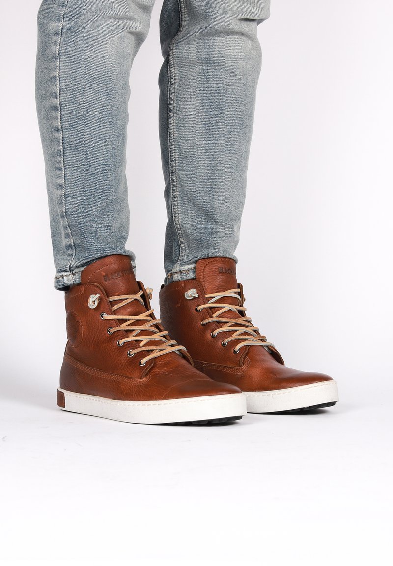 Zapatos de cuero marrón tipo bota alta con cordones beige, superficie texturizada, suelas de goma blanca y marca en la lengua y el costado.