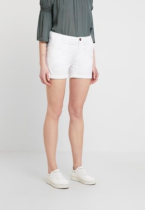 Frau trägt weiße Shorts, weiße Sneakers und eine grüne Bluse mit langen Ärmeln und steht vor einem schlichten weißen Hintergrund.