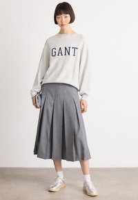 Svetlo šedá mikina s logom "GANT" v tmavomodrom, kombinovaná so šedou plisovanou midi sukňou. Model má na sebe strieborné tenisky a biele ponožky.
