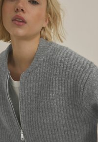 Grå ribbad zip-up tröja med hög krage, som har en strukturerad kläde och silverfärgad dragkedja. Under finns en vit topp delvis synlig.