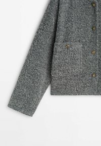 Veste grise texturée avec des fermetures à boutons et une poche avant à rabat dotée de quincaillerie en laiton. Le tissu a une apparence douce et duveteuse.