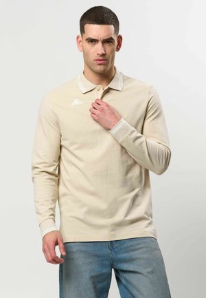 Mann trägt beiges Langarm-Poloshirt mit weißem Logo auf der Brust und hellblaue Jeans, steht vor einem schlichten, hellen Hintergrund.