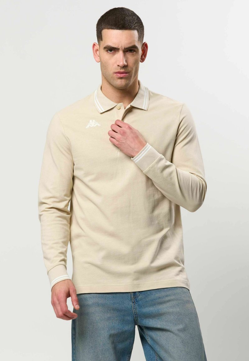 Homme portant un polo beige à manches longues avec un logo blanc sur la poitrine et un jean bleu clair, debout devant un fond clair uni.