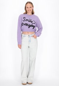Paarse cropped sweater met zwarte tekst "Smile everyday," gecombineerd met lichtgewassen flared jeans en witte sneakers. Glad brei textuur.