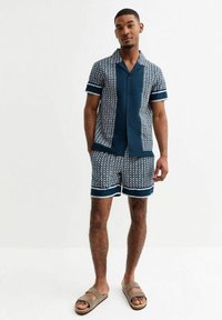 Short-sleeve, dark blue button-up shirt with a geometric white pattern, paired with matching shorts and tan sandals. Cotton fabric, relaxed fit.