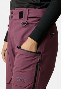 Bordeauxfarbene Outdoor-Hosen mit einer figurbetonten Passform, ausgestattet mit Reißverschlusstaschen, Gürtelschlaufen und einem Logo auf dem Oberschenkel. Hergestellt aus strapazierfähigem, wetterfestem Material.