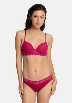 Vrouw in bijpassende donkerroze beha en met kant afgewerkte slip, staand tegen een egale witte achtergrond.
