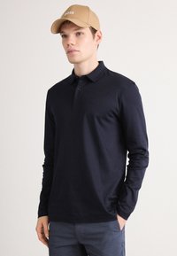 Marineblaues Langarm-Polohemd mit klassischem Kragen, Deux-Button-Leiste und dezenter Logo-Applikation auf der Brust. Begleitet von einer beigen Kappe.