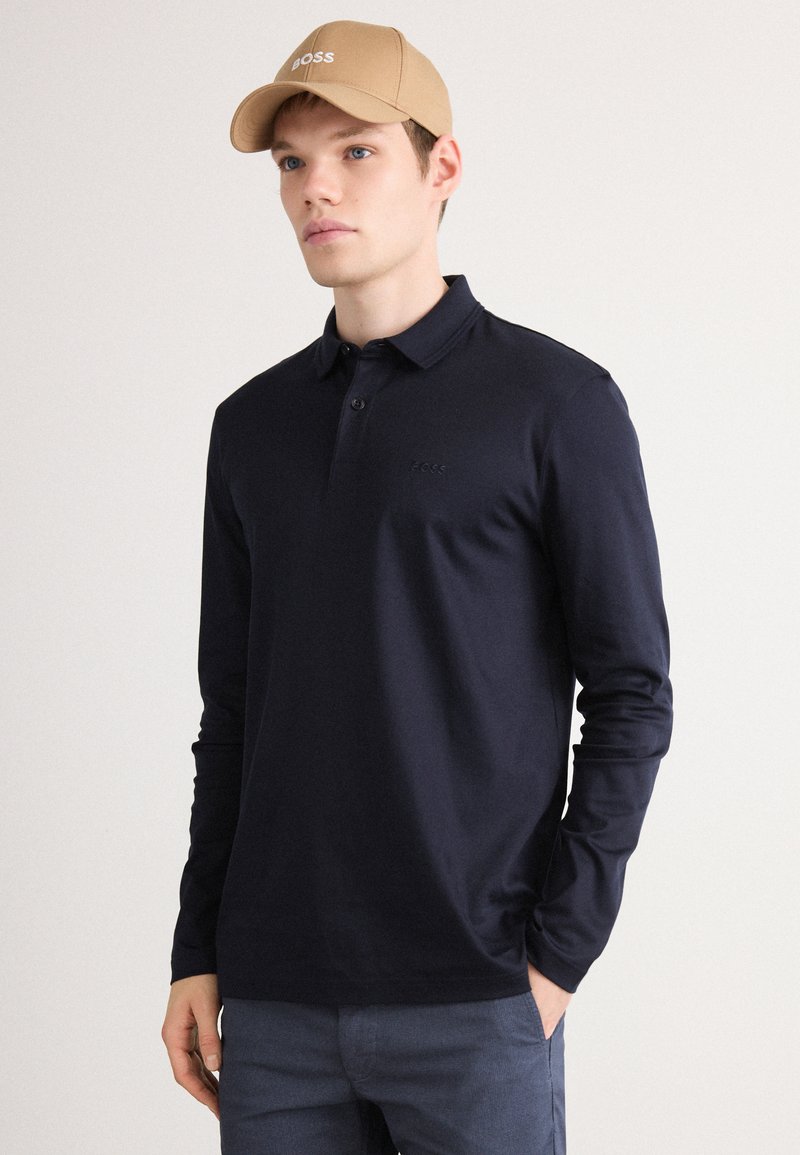 Marineblaues Langarm-Polohemd mit klassischem Kragen, Deux-Button-Leiste und dezenter Logo-Applikation auf der Brust. Begleitet von einer beigen Kappe.