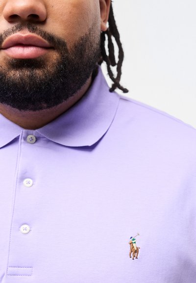 Κοντινό πλάνο ενός άνδρα με γένια που φοράει ανοιχτό μοβ πόλο πουκάμισο με κεντημένο λογότυπο παίκτη πόλο κοντά στο στήθος.