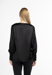 Schwarze Satinbluse mit langen Ärmeln, lockerem Schnitt, dezentem Glanz und gerafften Manschetten; hat einen runden Ausschnitt und eine glatte Textur.
