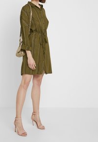 Robe boutonnière vert olive avec des rayures verticales et une ceinture à nouer, présentant des manches longues. Portée avec des escarpins beiges à lanières et un petit sac.