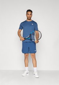 Blaues Sport-T-Shirt und Shorts, aus leichtem Stoff gefertigt. Ein Schläger wird in einer Hand gehalten. Weiße Sportschuhe und Socken vervollständigen das Outfit.