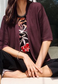 Žena sedící v tureckém sedu, oblečená v bordovém žebrovaném kardiganu přes květinové tričko, černých kalhotách a stříbrném náramku na levém zápěstí.