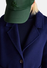 Cappotto di lana blu navy con revers a punta e un bottone marrone, indossato sopra un maglione scuro, abbinato a un berretto verde con logo.