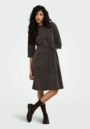 Une femme porte une robe marron foncé ceinturée et boutonnée sur le devant, des chaussettes à motifs et des chaussures noires, sur un fond clair uni.