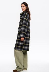 Abrigo de lana gris a cuadros con acentos en verde y amarillo, que presenta un alto cuello y puños abotonados, combinado con pantalones holgados de color verde claro.