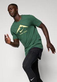 T-shirt Nike Trail vert avec une insigne de montagne jaune, porté avec des leggings noirs, mettant en avant un design sportif et un ajustement flexible.