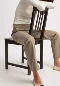 Beige tailored pants with a smooth texture, yhdistetään valkoiseen tiiviiseen ribbitoppiin, ja ne on paritettu kermaisten balettikenkien kanssa, istuen puisella tuolilla.