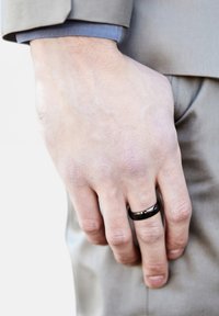 Sort metalring på en hånd med lys hudtone, iført en lysegrå dragt med en blå manchet synlig. Hånden er placeret nær taljen.